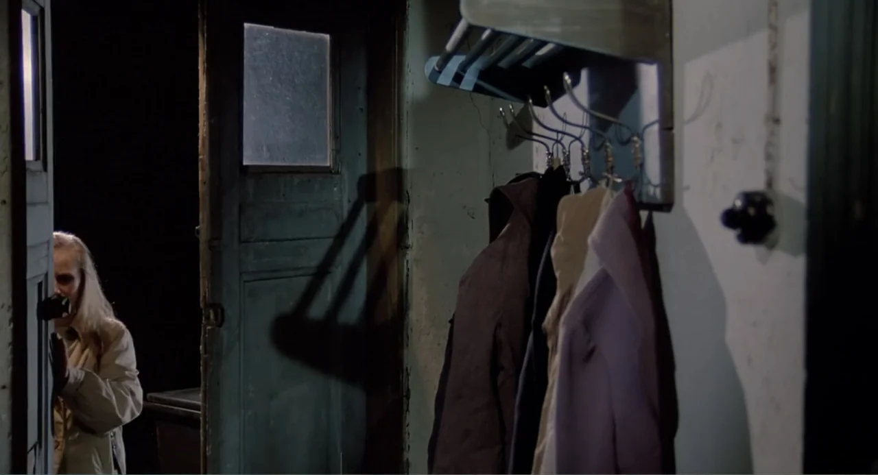 The Match Factory Girl (1990) movie still: medium — A dimly lit hallway features a coat rack holding several jackets on the wall. A woman with long…