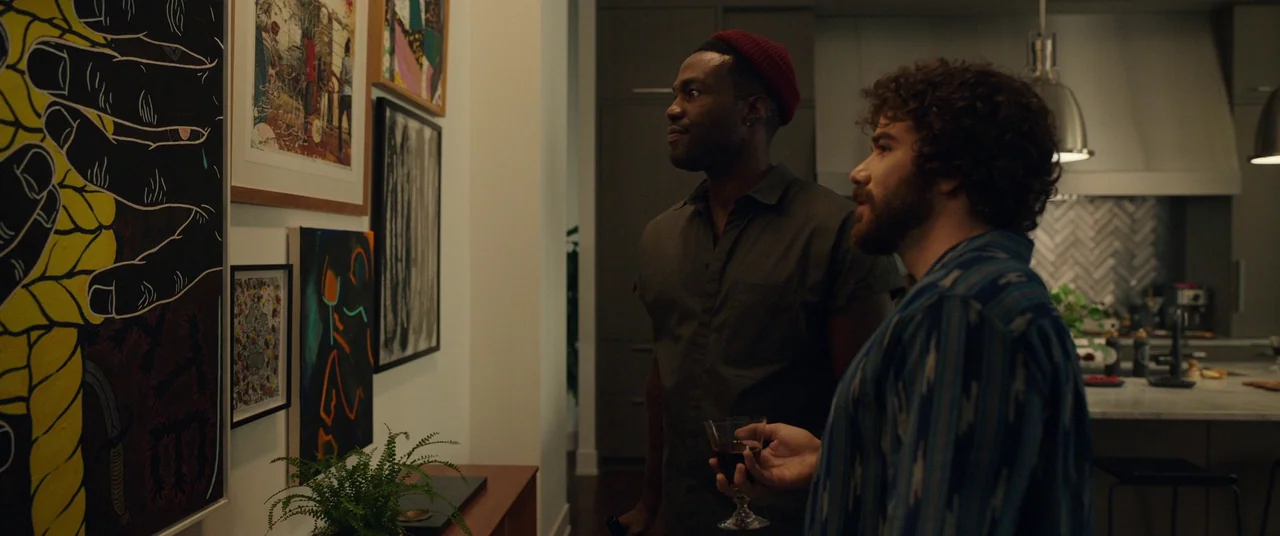 Candyman (2021) movie still: medium — Two men stand in a modern apartment hallway filled with framed art pieces. They hold glasses of red wine while…