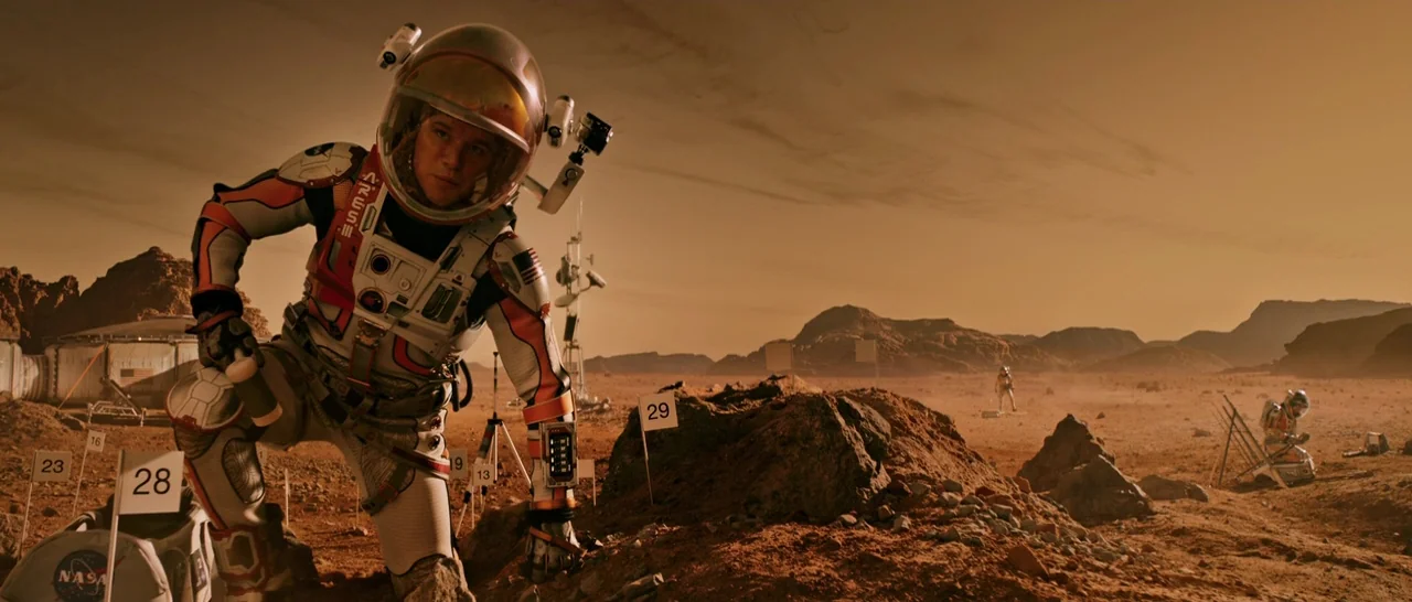 The Martian (2015) movie still: fullbody — The surface of Mars features reddish rock, dust, and numerous numbered surveying flags. An astronaut…