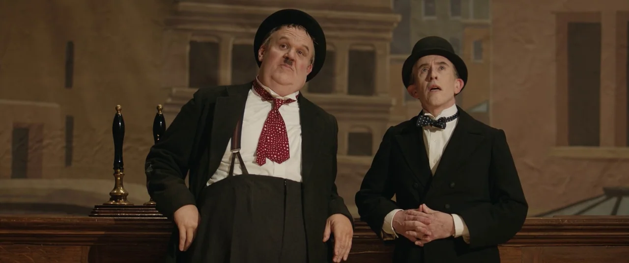 Domino (2005) movie still: medium — A theatrical stage set depicting a pub interior with a painted backdrop of buildings. Two men in bowler hats and…