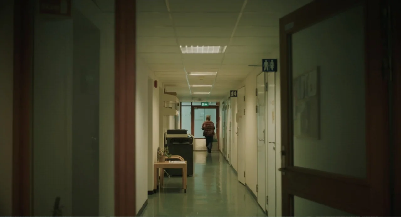 Ninjababy (2021) movie still: wide — A long, brightly lit hospital hallway is flanked by white doors and signs. A woman in a patterned sweater walks…