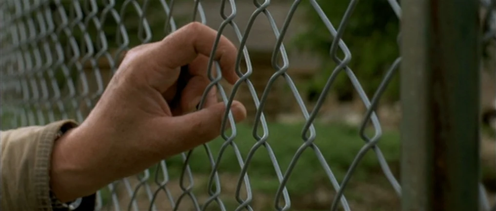 The Intruder (2004) movie still: closeup — A close-up shot focuses on a human hand holding a grey chain-link fence. The background shows blurred…