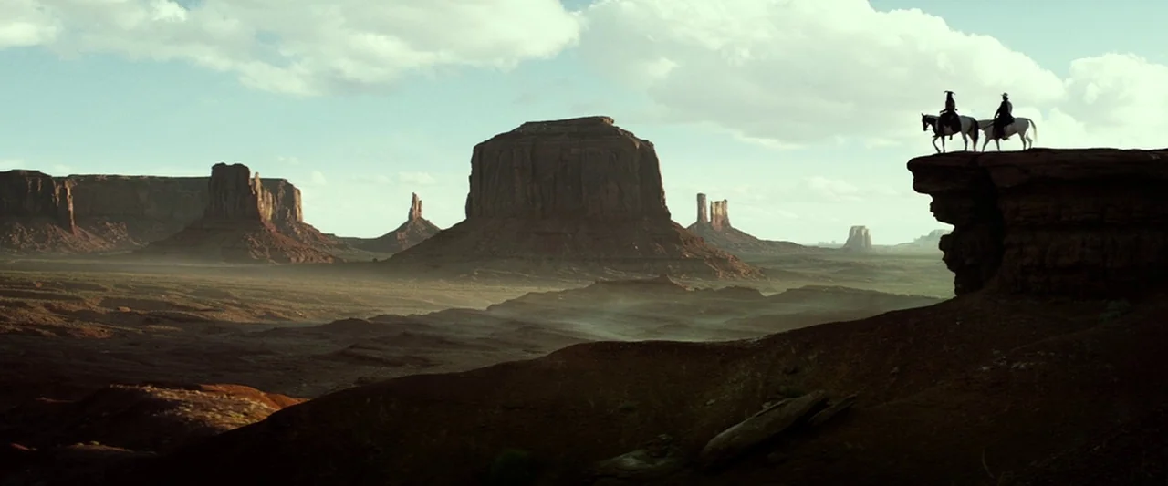 The Lone Ranger (2013) movie still: wide — The arid expanse of Monument Valley features towering sandstone mesas under a bright sky. Two riders…