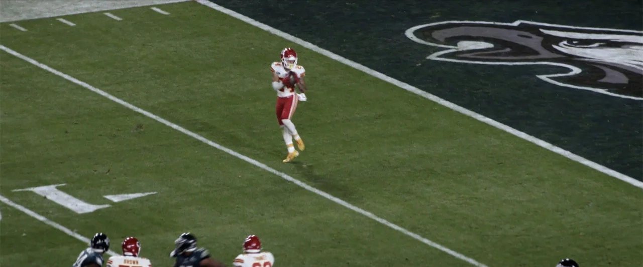Kelce (2023) movie still: wide — A professional football field displays white yard lines and a large team logo emblem. A player in a white uniform…
