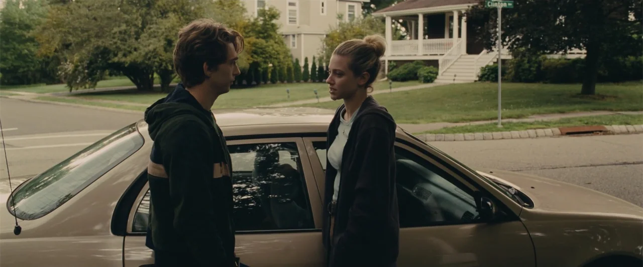 Chemical Hearts (2020) movie still: medium — A tan sedan is parked on a suburban road in front of a house. Two young adults stand beside the vehicle…