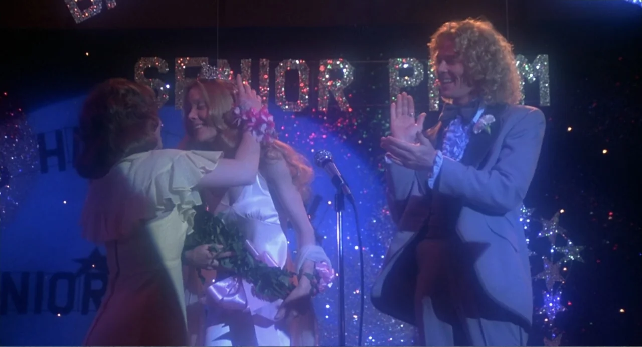 Carrie (1976) movie still: medium — A dark gymnasium decorated with sparkling star cutouts features a stage for a dance. Two girls and a boy stand…