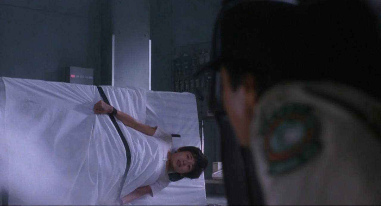 Godzilla vs. SpaceGodzilla (1994) movie still: medium — A dimly lit room features a patient restrained to a medical bed under soft light. A figure…