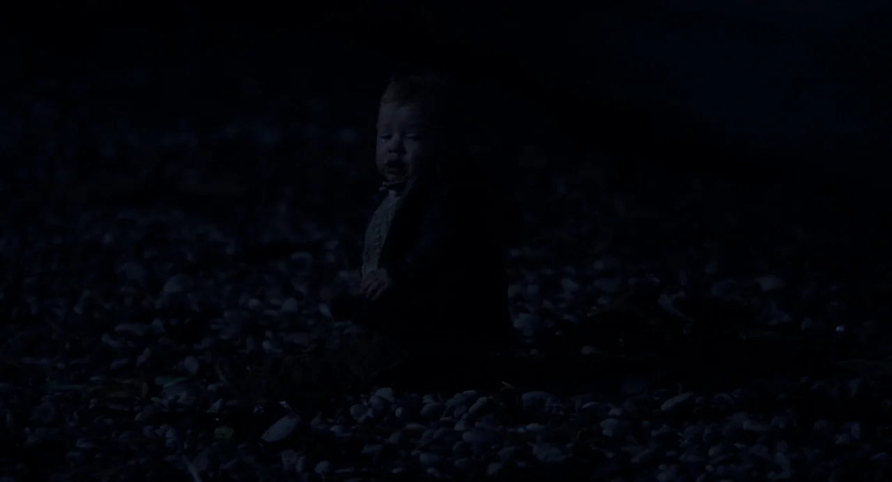 Under The Skin (2013) movie still: medium — A dark pebble beach at night with the ground covered in smooth stones. A young child sits alone on the…