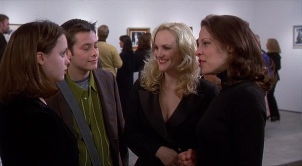 Pecker movie still: medium — An interior gallery space with white walls and framed artwork in the background. Two men and two women are gathered…