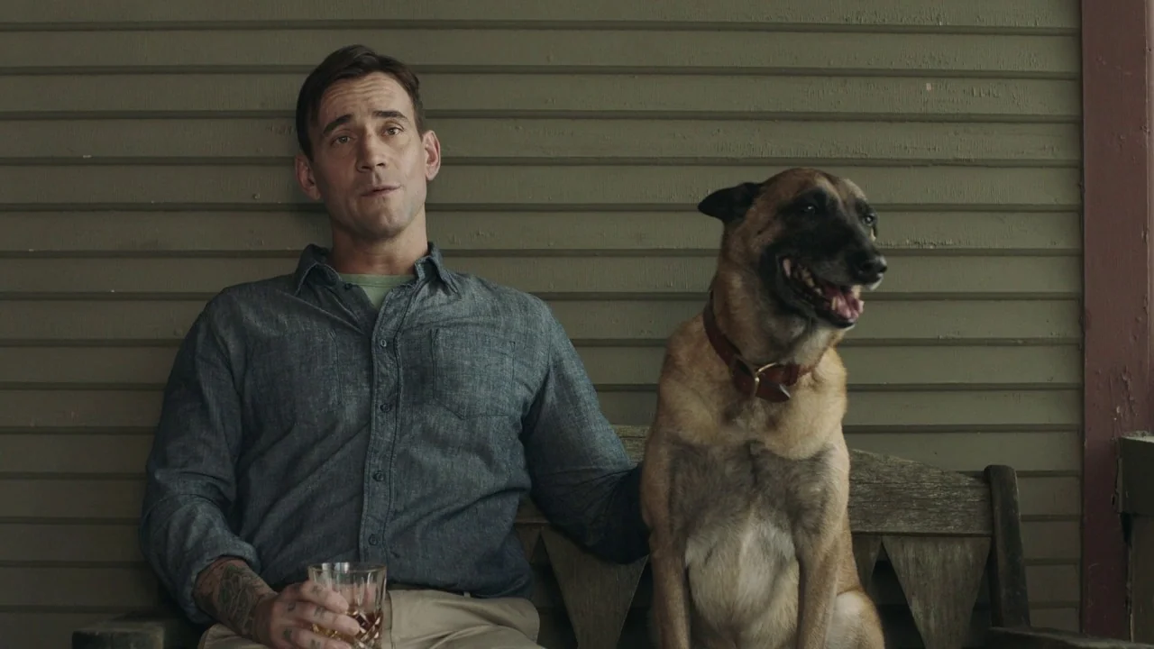 Girl on the Third Floor (2019) movie still: medium — A man and his dog sit on a wooden bench against olive green wood siding. The man holds a glass…