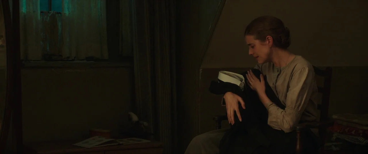 Sunset Song (2015) movie still: medium — A dimly lit room with a wooden chair and a small table near a window. A young woman sits in the chair while…