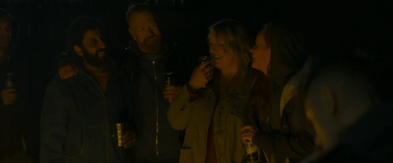 Ali & Ava (2021) movie still: medium — A group of friends stands together outdoors at night near a warm, unseen fire. They are talking and drinking…