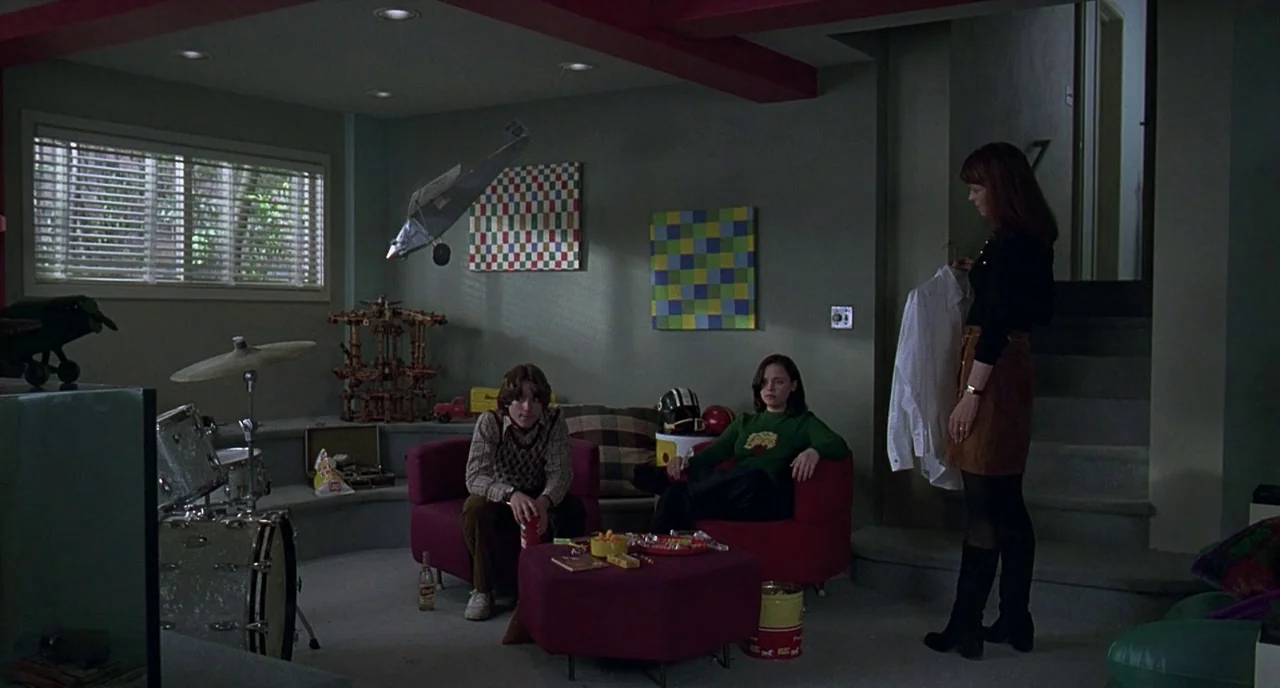 The Ice Storm (1997) movie still: wide — A basement room features red circular chairs, geometric art, and a drum kit. A young man and woman sit…