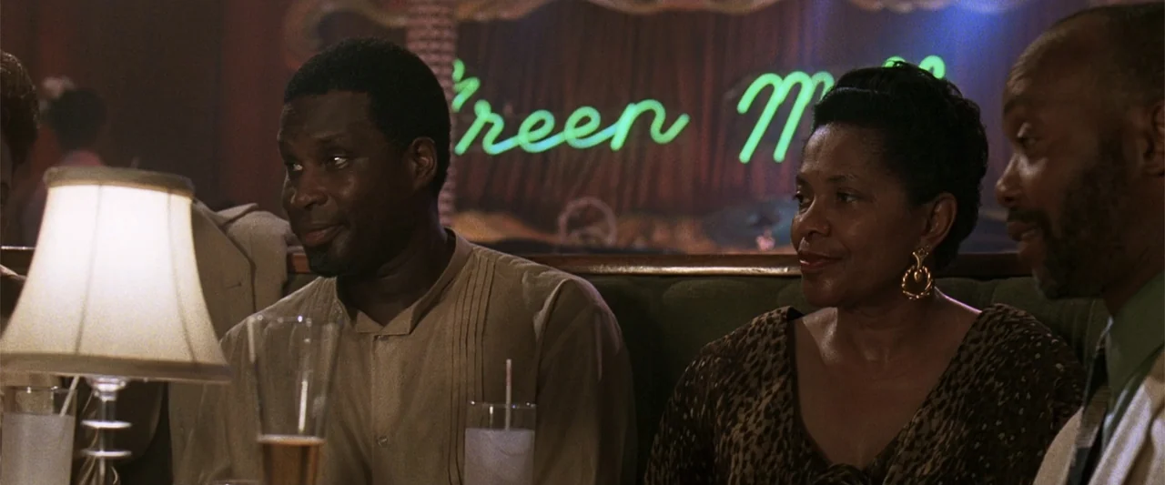 A Family Thing (1996) movie still: medium — A dark booth at a nightclub with a glowing neon sign in the background. Three people sit at a table…