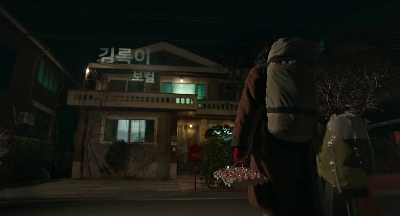 Microhabitat (2017) movie still: fullbody — A suburban street at night features a two-story house with illuminated windows and signage. A person…
