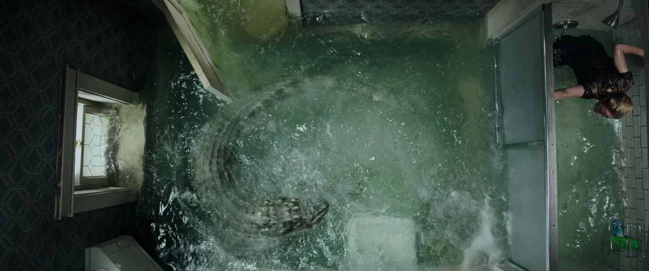 Crawl (2019) movie still: wide — A flooded room contains green murky water and a large thrashing alligator. A woman floats in the water near the…