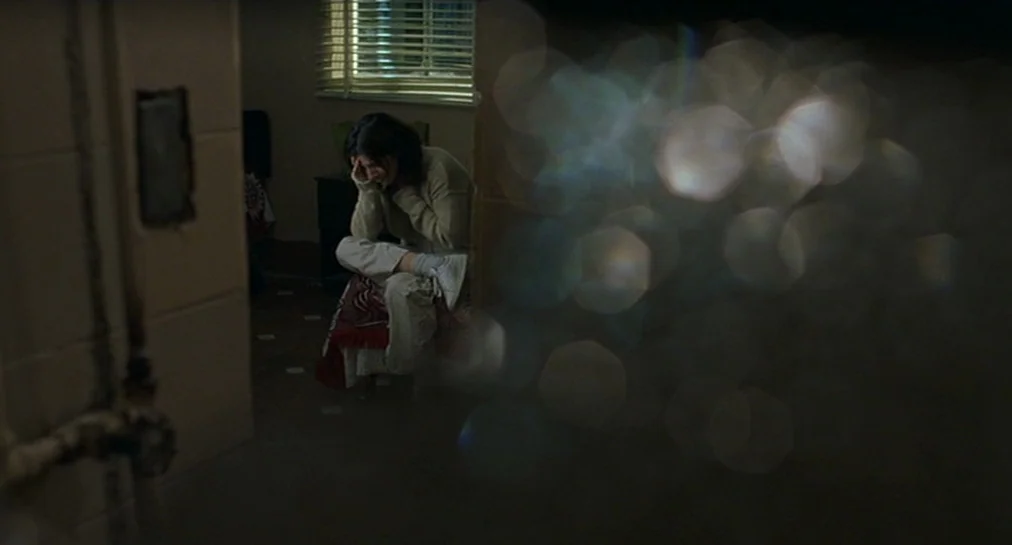 Y Tu Mamá También (2001) movie still: wide — A dimly lit room features a person sitting on an elevated tiled platform. The subject rests their head…