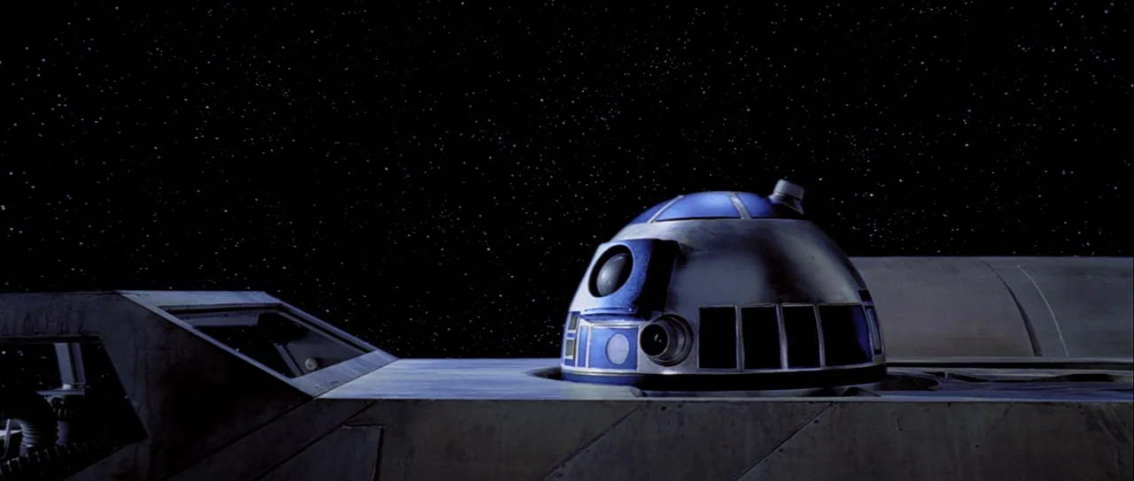 The Empire Strikes Back (1980) movie still: medium — A silver and blue astromech droid sits in the circular slot of a metallic ship. The vastness of…