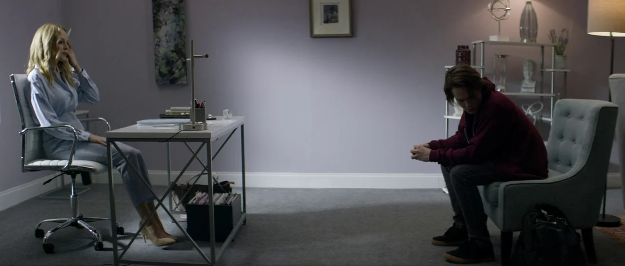 Suitable Flesh (2023) movie still: wide — A spare, modern room contains a desk and two chairs separated by open floor space. A woman in business…