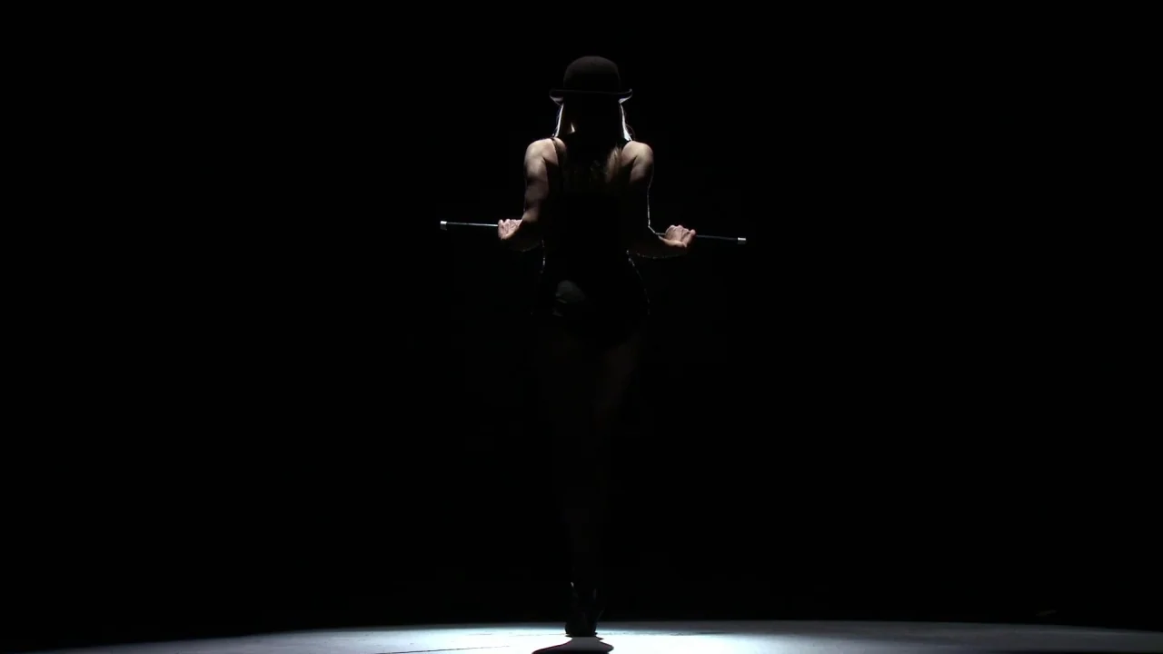 Electra Luxx (2010) movie still: fullbody — A dark, empty space is illuminated by a single focused light from above. A woman in a leotard and bowler…