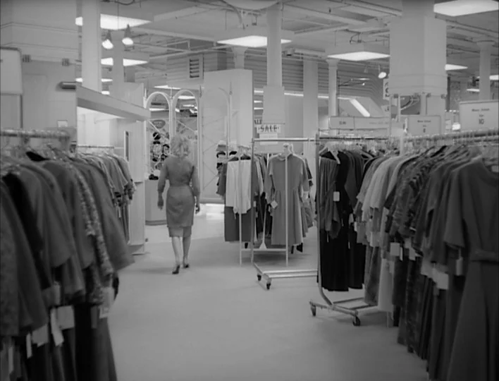 Carnival of Souls (1962) movie still: fullbody — A mid-century retail space filled with rows of rolling garment racks displaying dresses. A woman…