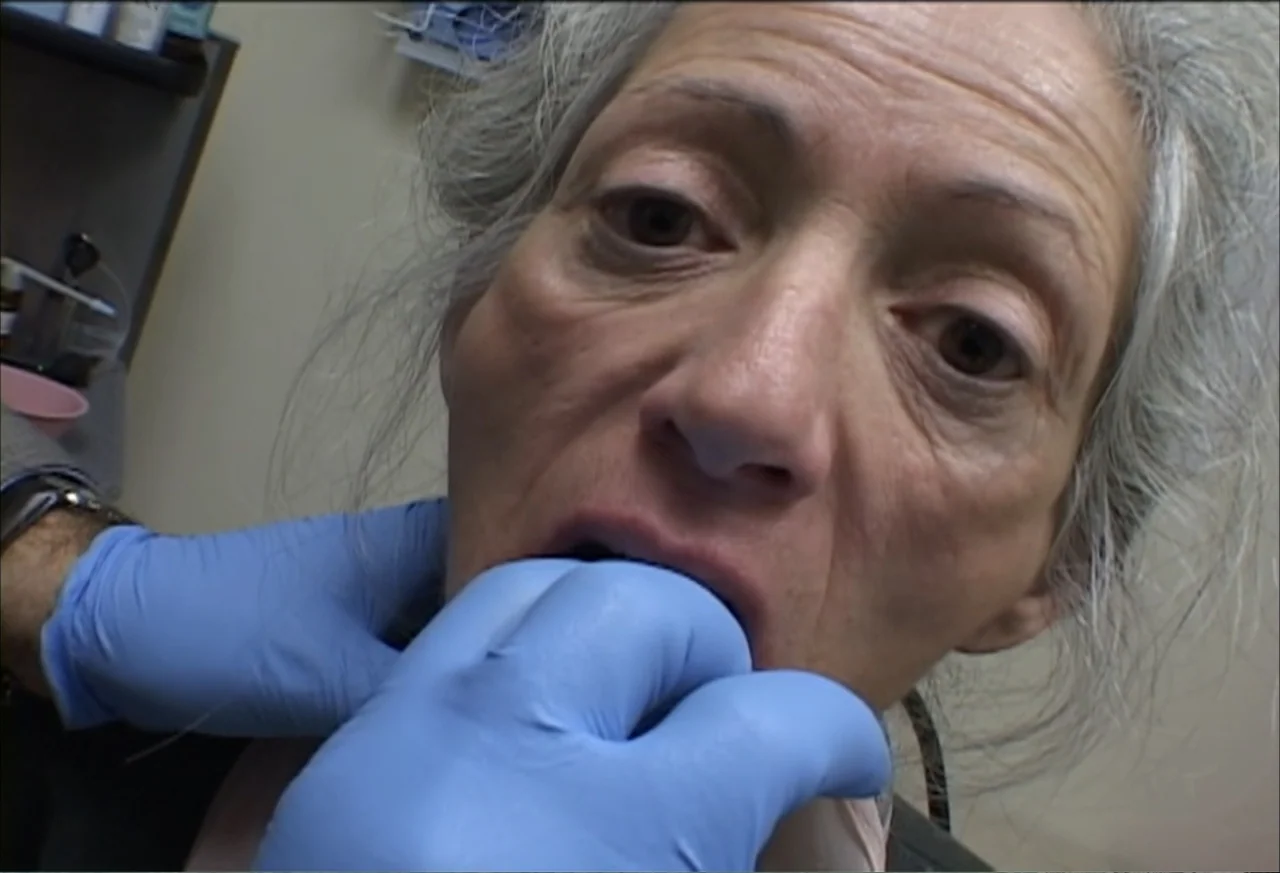 Plastic Disasters (2006) movie still: closeup — A clinical examination room with medical supplies blurred in the background. A doctor wears blue…