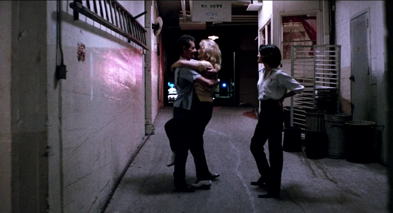 Desert Hearts (1985) movie still: fullbody — A narrow service corridor with concrete walls and industrial metal storage shelving. A man and woman…