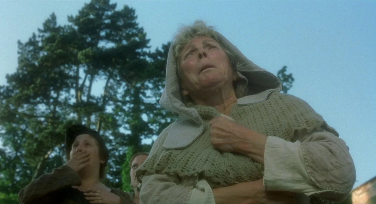 Quills (2000) movie still: medium — A group of women stand in an outdoor setting beneath a clear blue sky. An older woman in a hooded shawl gazes…