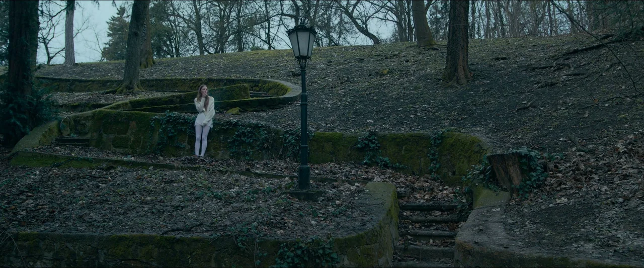Birds of Paradise (2021) movie still: wide — A quiet, mossy public space features a stone retaining wall and a vintage lamppost. A young woman in…