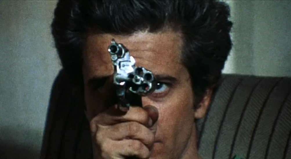 The Last House on the Left (2009) movie still: closeup — An interior space is dimly lit in a low-key fashion. A man points a revolver directly…