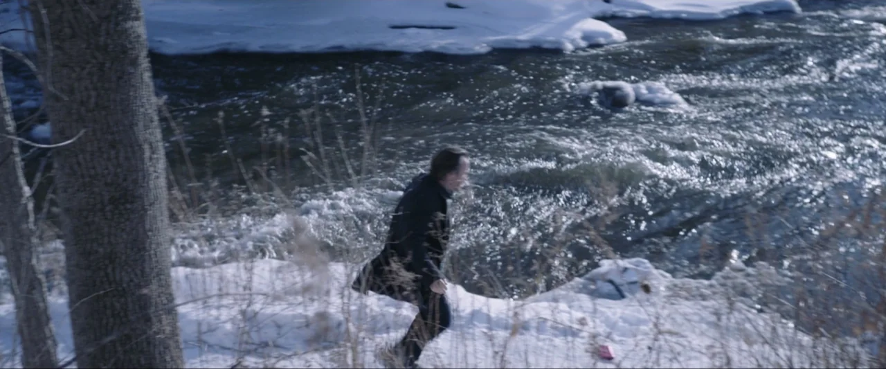 The Lie (2018) movie still: medium — A snowy riverbank features dry vegetation and a fast-moving, dark water stream. A man wearing a dark coat walks…