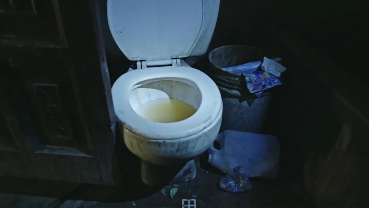 Finding the Way Home (1991) movie still: medium — A dimly lit, cramped restroom space features a stained white toilet bowl. Next to the toilet sits…