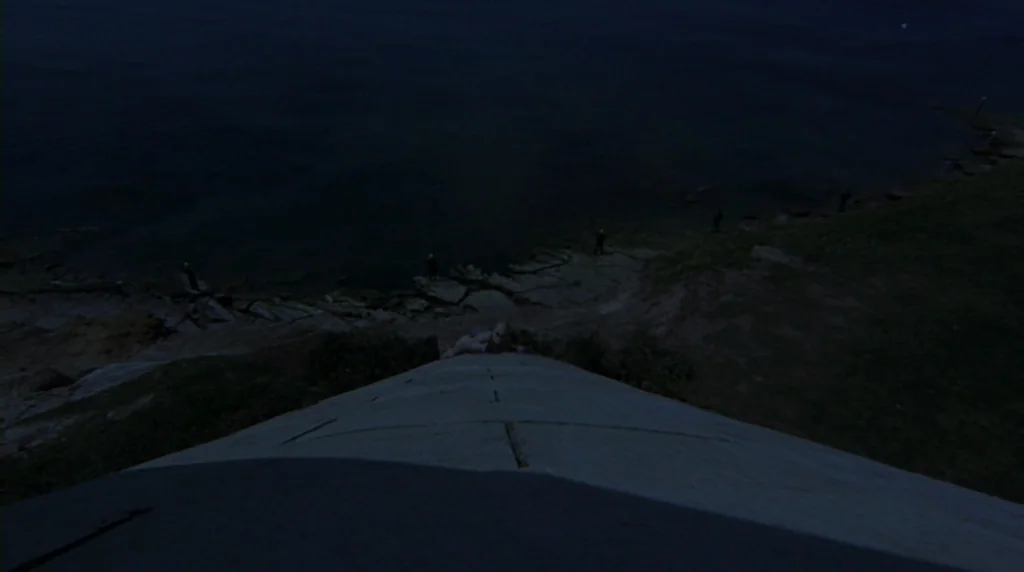 Danger Diabolik (1968) movie still: establishing — A dark, elevated concrete surface descends toward a jagged rocky shoreline and calm water. Small,…