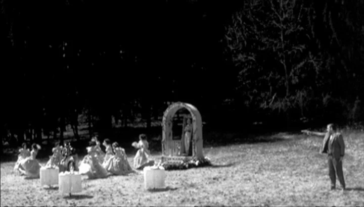 The American Astronaut (2001) movie still: wide — A group of women in light-colored dresses gathers near a small outdoor shrine in a field. A man in…