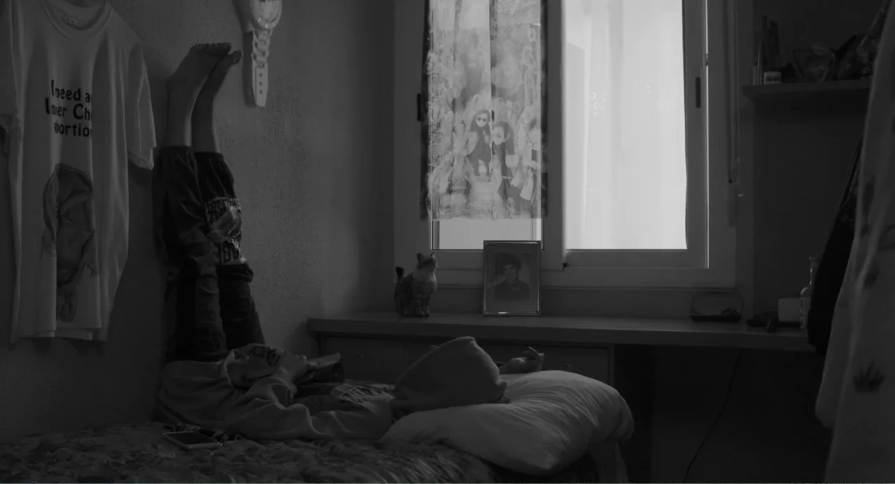 El Planeta (2021) movie still: medium — A cluttered bedroom with a single bed positioned next to a window. A person lies on their back with their…