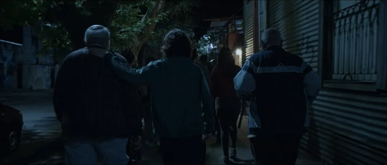 Gunman (2025) movie still: medium — A dark urban street at night with corrugated metal walls and overhanging trees. A small group of three people…