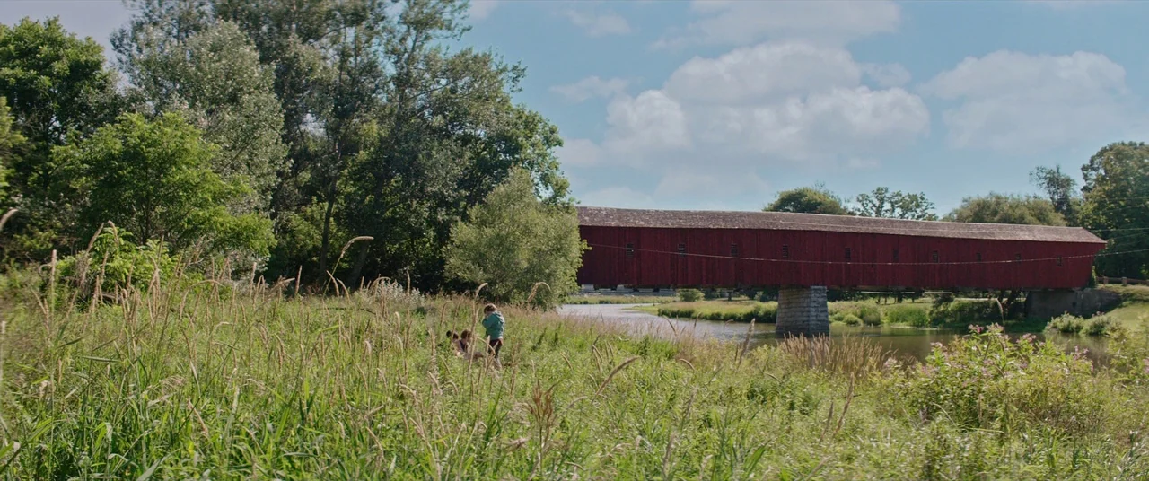 It (2017) movie still: wide — A tall, red-painted covered bridge spans a slow-moving river surrounded by dense summer foliage. Two small figures…