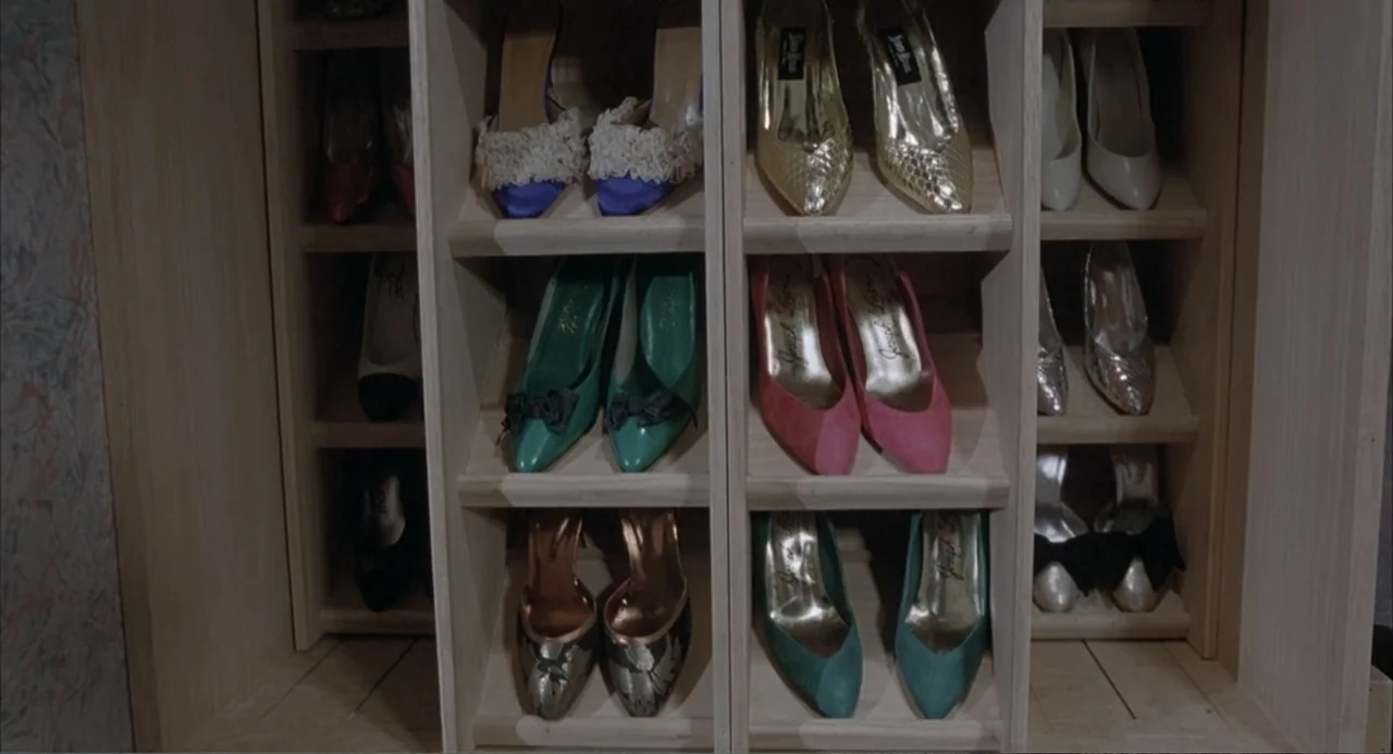 Overboard (1987) movie still: closeup — A wooden shelving unit displays multiple pairs of colorful women's high-heeled pumps. The shoes are arranged…