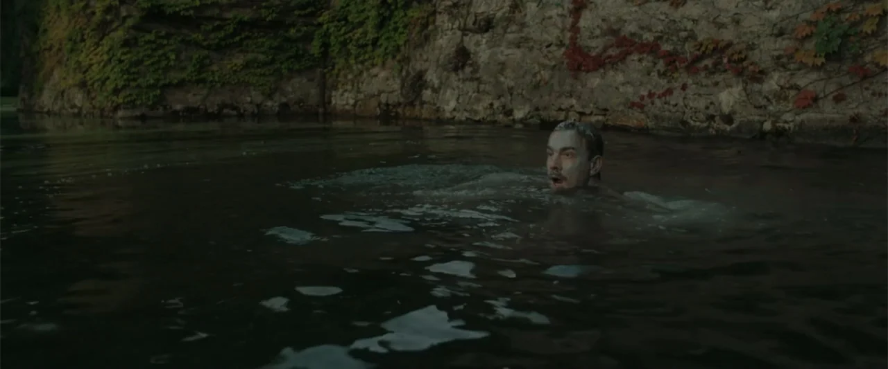 Peacock (2010) movie still: medium — A man swims in a murky river beside a mossy, ivy-covered stone wall. The subject keeps his head above water…