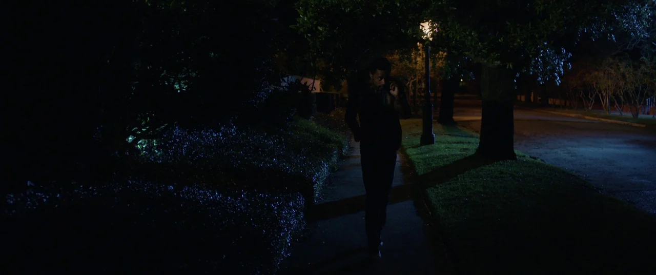 Get Out (2017) movie still: wide — A dark residential sidewalk at night is bordered by large trees and hedges. A man walks toward the camera while…