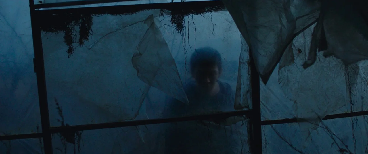 Burning (2018) movie still: medium — A dilapidated greenhouse structure with torn plastic panels blowing in the wind. A young person stands behind…