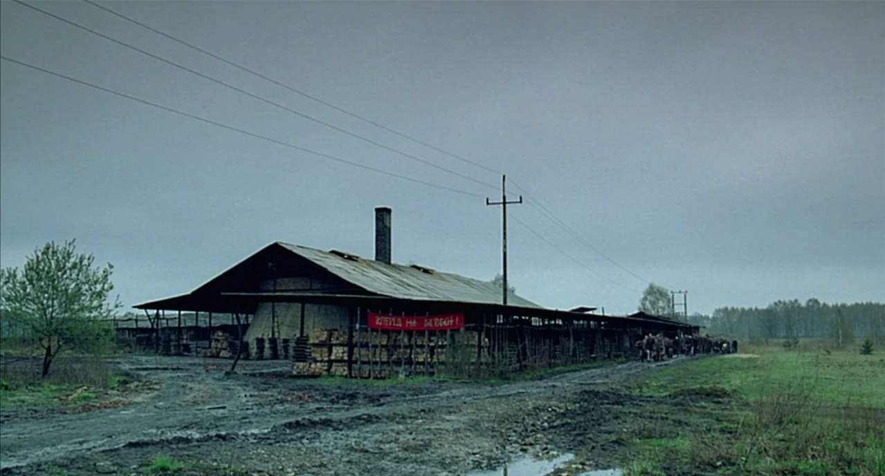 The Pianist (2002) movie still: wide — A rural industrial brickyard under a grey overcast sky features a long wooden shed. A red banner hangs across…