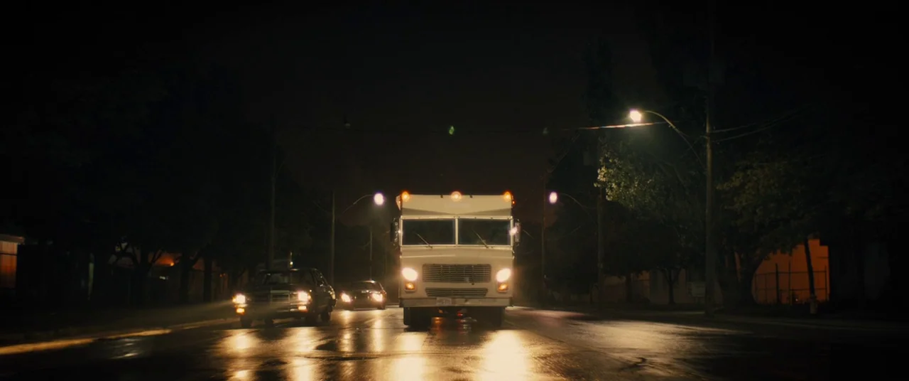 Free Fire (2016) movie still: wide — A dark night scene on a wet asphalt street lined with trees and poles. A white truck and a sedan approach the…