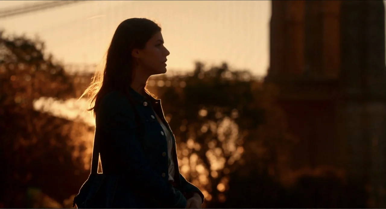 Can You Keep a Secret? (2019) movie still: medium — A woman stands in profile against the blurred backdrop of a bridge structure. She gazes out into…