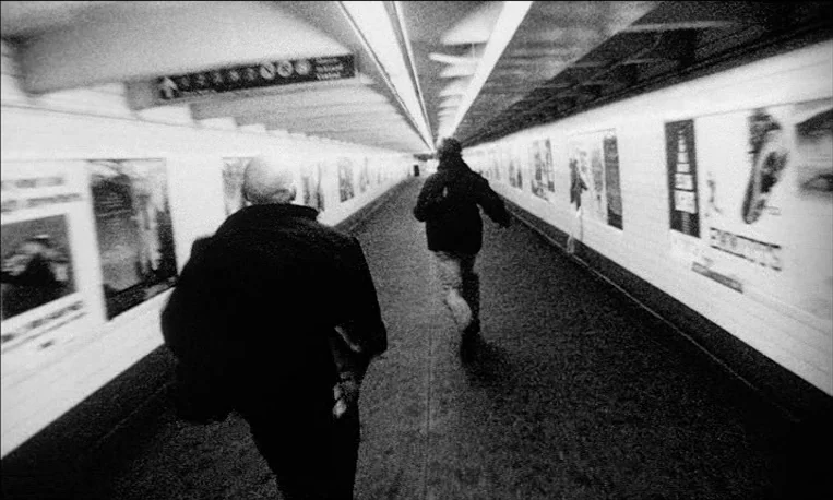 Pi (1998) movie still: medium — A long underground subway walkway with white tiled walls lined with advertisements. Two men run away from the camera…