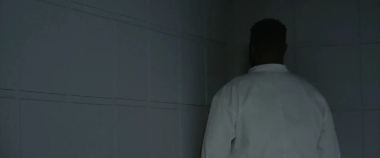 A Scam Called Love (2025) movie still: medium — A dim room features walls covered in square white tiles with distinct grout lines. A man wearing a…