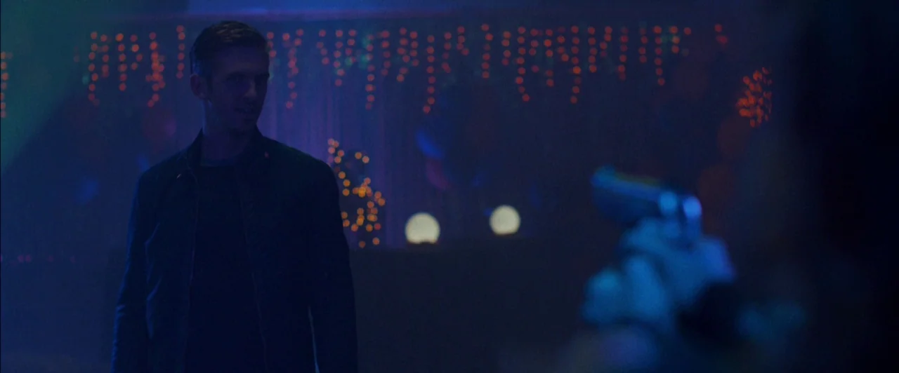 The Guest (2014) movie still: medium — A dimly lit room decorated with blurred strings of orange festive lights. A man stands in the blue-lit…