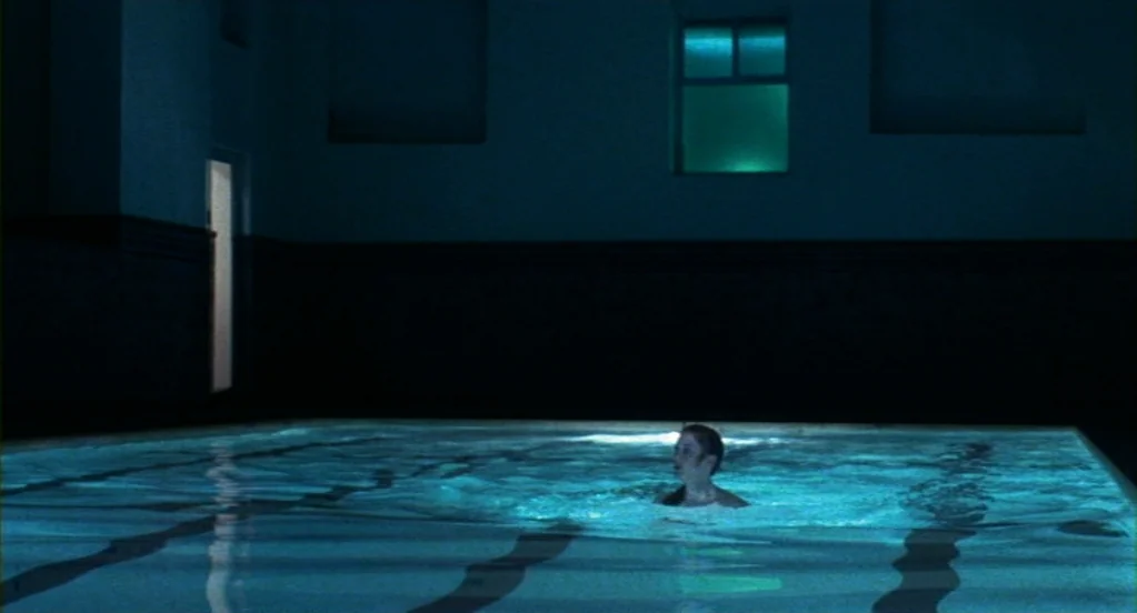 Cat People (1982) movie still: wide — An indoor swimming pool with dark walls and underwater lighting at night. A lone woman stands in the middle of…