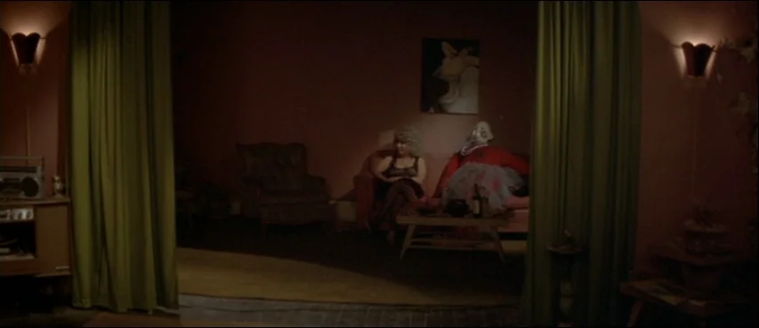 Blue Velvet (1986) movie still: wide — A dimly lit room features pink walls and green floor-to-ceiling curtains on both sides. Two women sit on a…