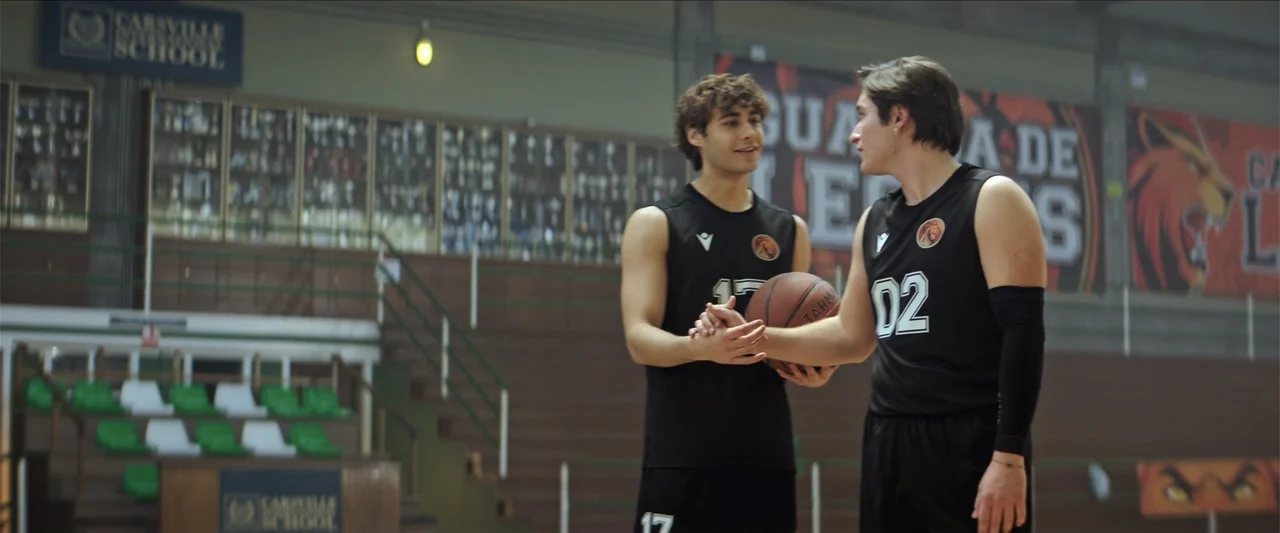 Tell Me Softly (2025) movie still: medium — A high school gymnasium interior features wooden bleachers and a large wall banner display. Two male…